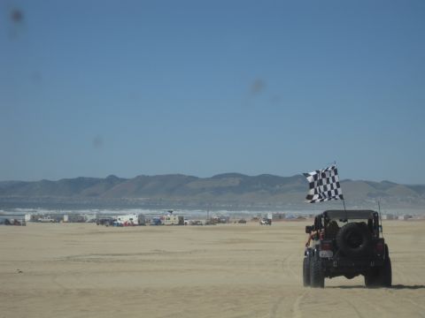 1989 Ford Bronco - A Jeep Wrangler flies a checkered flag while driving on a sandy beach with other vehicles and campers in the background.