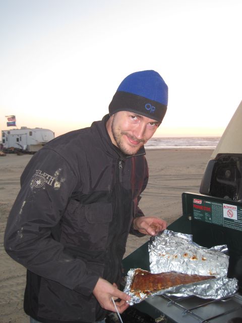 1989 Ford Bronco - A man cooks ribs on a portable grill on a beach, with an RV and flag visible in the distance.