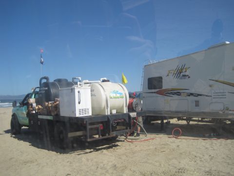 1989 Ford Bronco - A truck equipped with multiple water tanks and a cooler is parked next to a trailer on a sandy beach under a clear blue sky.