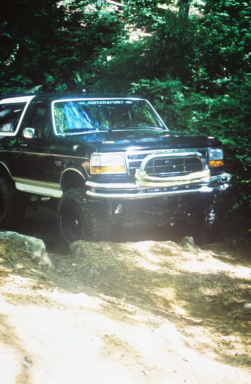 Black lifted full-size Ford Bronco crawling over rock on a wooded dirt trail