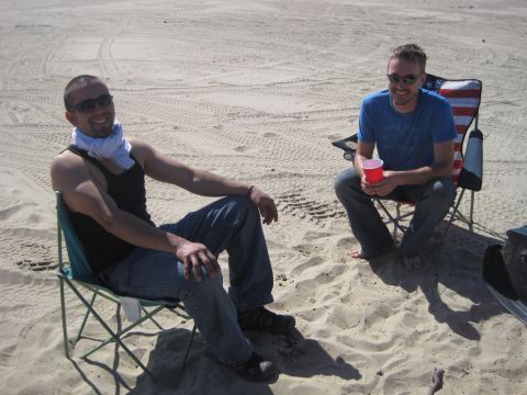 1989 Ford Bronco - Two men sit on camping chairs on a sandy beach, one holding a red cup.