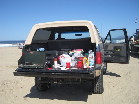 1989 Ford Bronco - A 1989 Ford Bronco is parked on the beach with its tailgate down, displaying picnic items and a portable stove.