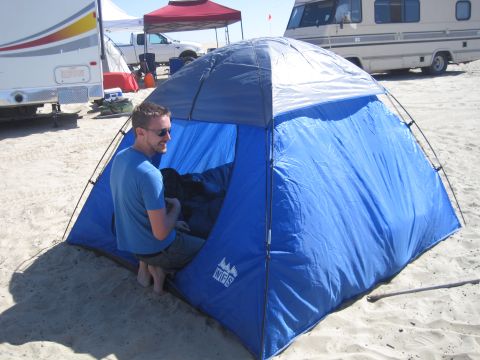 1989 Ford Bronco - A man sits in a blue tent on a sandy beach, with an RV and truck visible in the background.