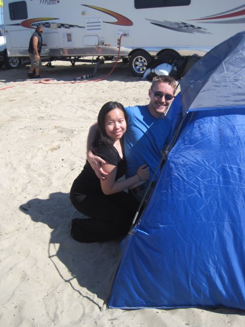 1989 Ford Bronco - A couple embraces near a tent on a sandy beach, with a trailer parked behind them.