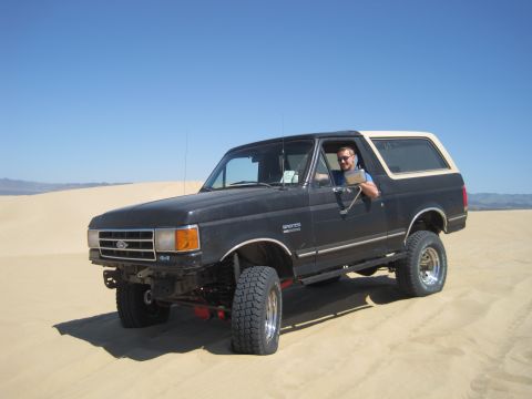 1989 Ford Bronco - A man drives a black Ford Bronco with a tan top through sand dunes under a clear blue sky.