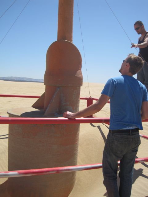 1989 Ford Bronco - Two men look up at a tall, orange, cylindrical structure in a desert landscape with sand dunes under a clear blue sky.