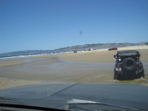 1989 Ford Bronco - Several vehicles are seen driving on a beach with the ocean and hills in the background.
