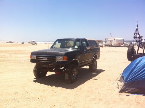 1989 Ford Bronco - A black 1989 Ford Bronco with a tan top is parked on a sandy beach with RVs and a tent in the background.