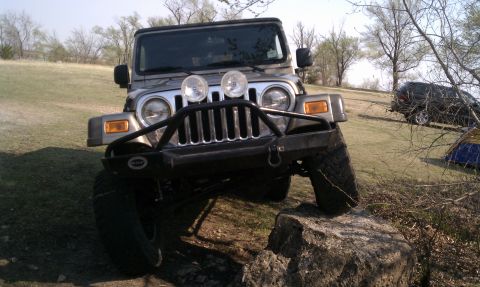 2005 Jeep Wrangler - This silver 2005 Jeep Wrangler is shown from the front as it maneuvers over rocks, with another vehicle and tents visible in the background.