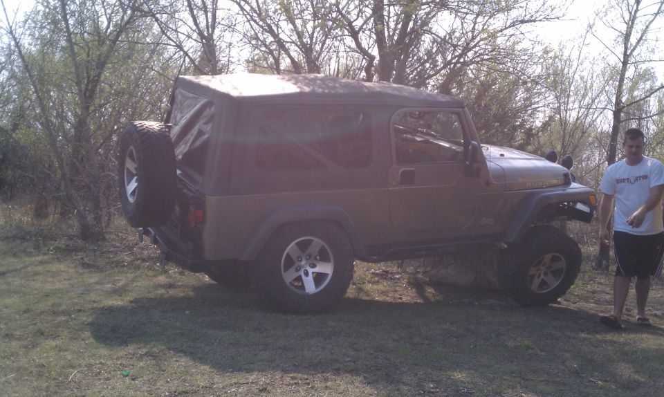 Brown Jeep Wrangler with a soft top, spare tire on the back, parked on grass with trees in the background
