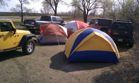 2005 Jeep Wrangler - A grassy campsite with three dome tents pitched in front of parked vehicles, including a yellow Jeep Wrangler JK Rubicon Unlimited at left,…