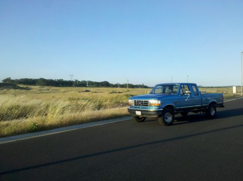 1992 Ford F-150 - A blue 1992 Ford F-150 pickup truck is seen driving on a road with a grassy field beside it.