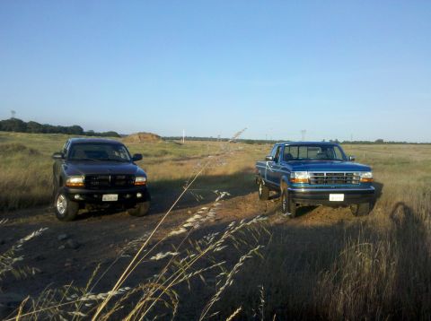 1992 Ford F-150 - A blue 1992 Ford F-150 and a dark colored truck are parked on a dirt road in a grassy field under a clear blue sky.