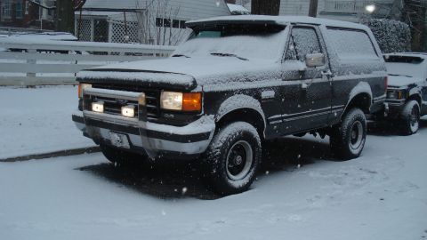 1988 Ford Bronco - A dark Ford Bronco sits parked on a snowy street, partially covered in snow.