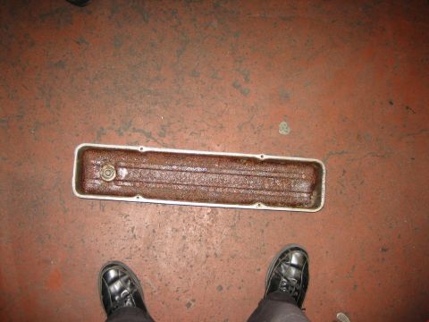 1998 Ford Crown Victoria - long metal valley cover full of rusty sludge lies on a red concrete floor between a person's black work boots.