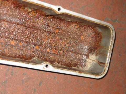 1998 Ford Crown Victoria - Macro shot of thick orange-brown rust and sludge caked inside a long sheet-metal engine cover on the floor.