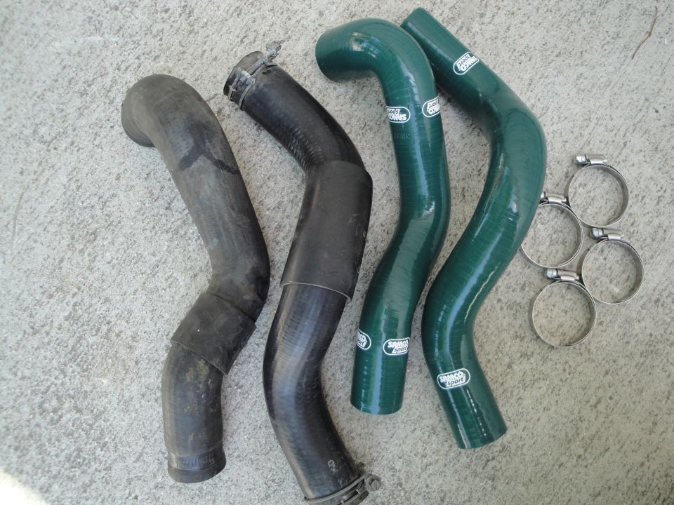 Two black rubber hoses and two green silicone hoses with clamps on a concrete surface. The green hoses have 'Samco Sport' branding