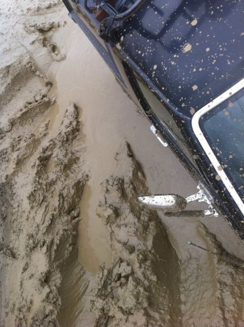 1990 Ford Bronco - A black SUV is deeply stuck in thick mud, with visible tire tracks and mud splattered across its body.