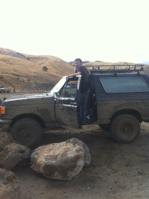 1990 Ford Bronco - A man stands in the open driver's door of a mud-covered 1990 Ford Bronco, parked outdoors with a roof rack.