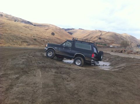 1990 Ford Bronco - A black Ford Bronco navigates a muddy terrain with hills in the background.