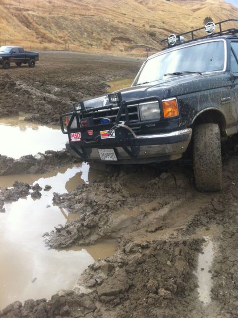 1990 Ford Bronco - This Ford F-Series truck is stuck in a muddy puddle, with a second truck visible in the background.