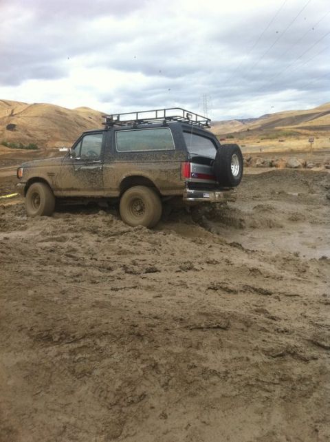 1990 Ford Bronco - Muddy 1990 Ford Bronco navigating rough terrain, equipped with a roof rack and external spare tire.