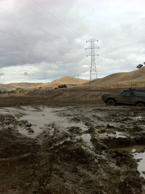 1990 Ford Bronco - A brown SUV is stuck in a muddy field with power lines and rolling hills in the background.