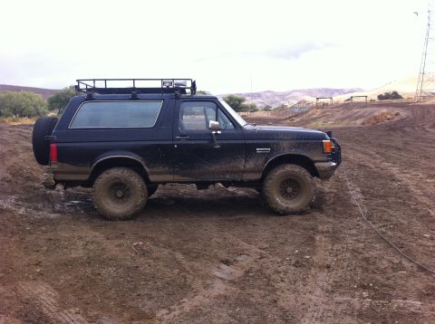 1990 Ford Bronco - This black 1990 Ford Bronco navigates a muddy off-road course, equipped with a roof rack and a rear-mounted spare tire.