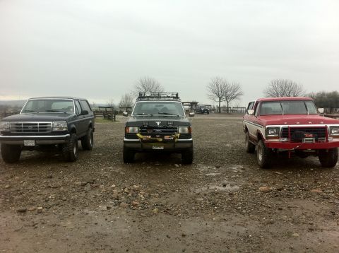 1990 Ford Bronco - Three Ford Broncos are parked in a gravel lot under an overcast sky.