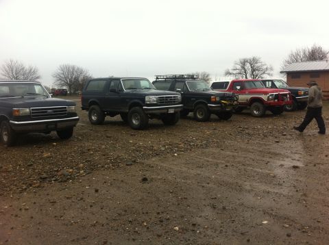 1990 Ford Bronco - Several Ford Broncos and trucks are parked in a gravel lot under an overcast sky.