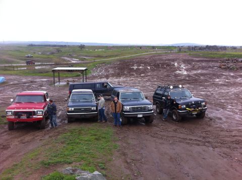 1990 Ford Bronco - A group of SUVs and a pickup truck are parked on a muddy field, with a scenic, overcast sky in the background.