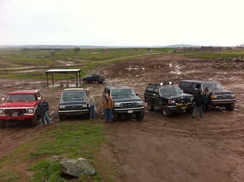 1990 Ford Bronco - A group of off-road vehicles are gathered on a muddy terrain under an overcast sky.