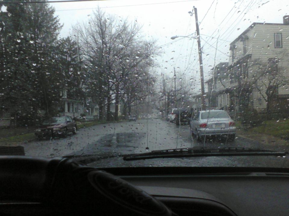 View from inside a car looking out at a rainy street with houses and parked cars