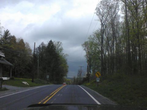 1988 Ford Bronco - A lone red car drives down a rural road under a cloudy sky.