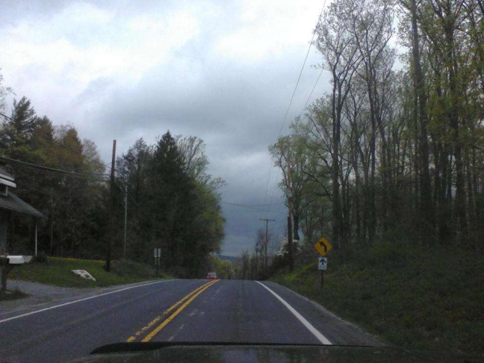 Red car seen from behind on a two-lane road with trees and a cloudy sky