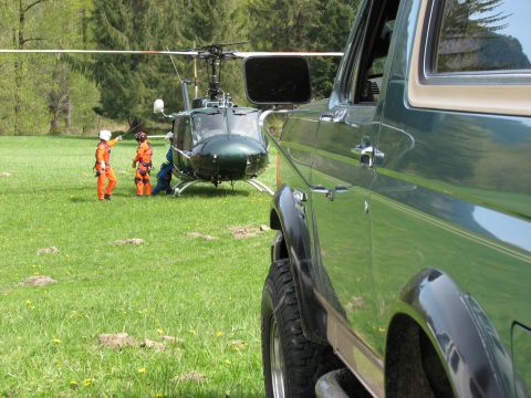 1990 Ford Bronco - A helicopter sits in a grassy field with two individuals in orange suits nearby, while the side of a green SUV is in the foreground.