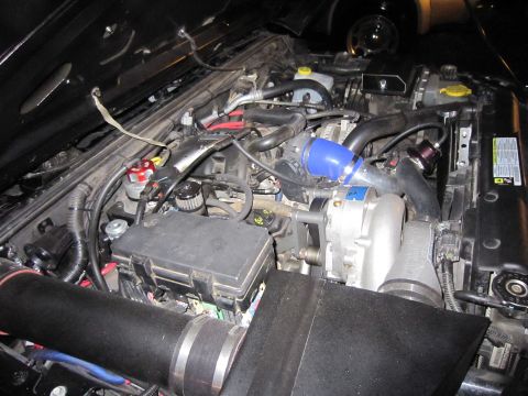 2008 Jeep Wrangler - Engine bay of a modified vehicle showing a belt-driven supercharger with a polished housing, a blue silicone elbow feeding the inlet, a pur…
