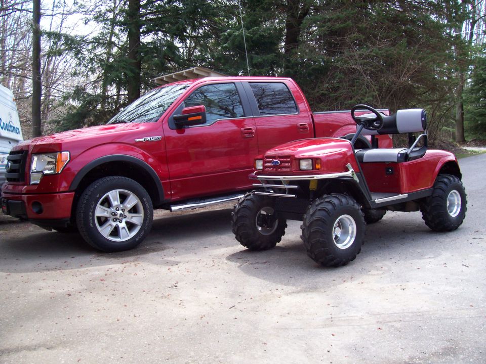 Red pickup truck parked next to a red golf cart with oversized tires. The truck appears to be a Ford F-150 from the 2009-2014 generation