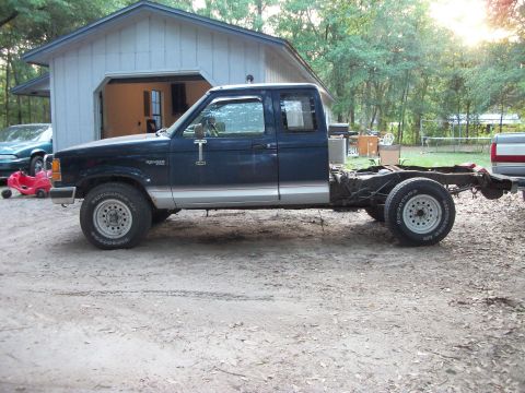 1994 Ford Explorer - A dark blue pickup truck chassis with a cab sits in a dirt lot next to a garage, with another vehicle partially visible in the background.