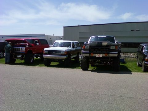 1989 Ford Bronco - Three lifted SUVs are parked in a line outside a building on a sunny day.