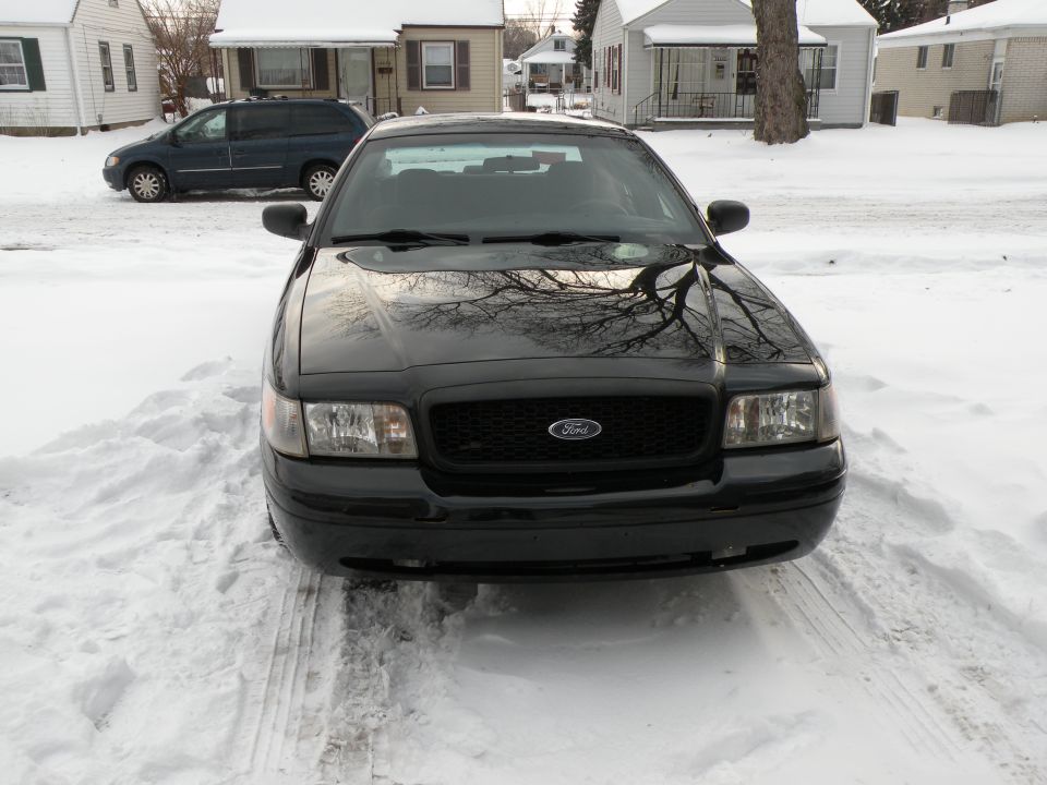 Black 2005 Ford Crown Victoria parked in snow, front view. A dark blue minivan is visible in the background