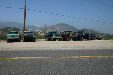 1991 Ford Bronco - A group of modified trucks are parked on a dirt lot with a mountainous backdrop under a clear blue sky.