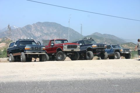 1991 Ford Bronco - A lineup of four modified off-road vehicles, including a 1991 Ford Bronco, are parked on a dusty lot with mountains in the background.