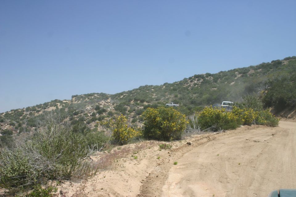 Two white SUVs driving up a dusty, winding dirt road on a hillside with green brush and yellow flowers