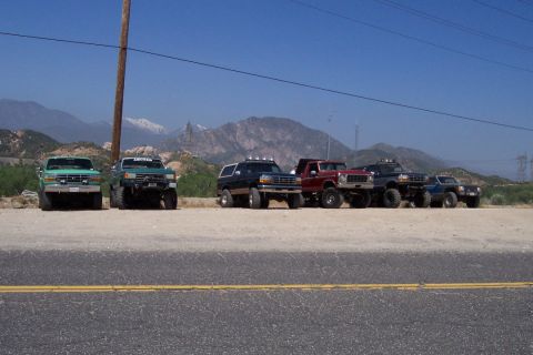 1995 Ford Bronco - A group of modified trucks and SUVs are parked on a dirt lot with a mountain range behind them.