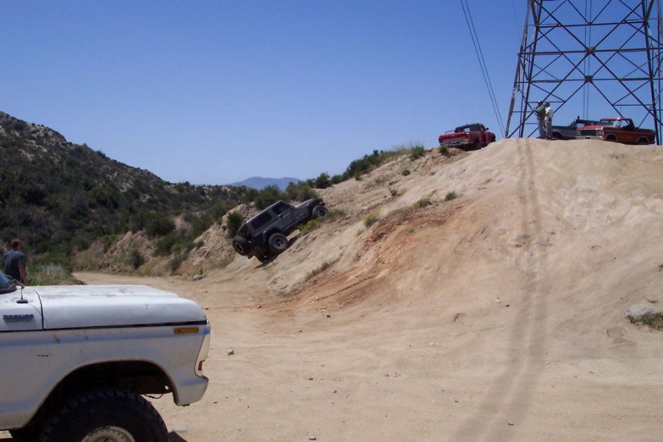 White truck in foreground, dark SUV climbing a steep dirt hill, two red trucks parked on top of the hill near a power line tower