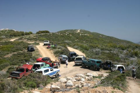 1991 Ford Bronco - Several off-road vehicles are gathered on a dusty trail with hills and brush in the background.