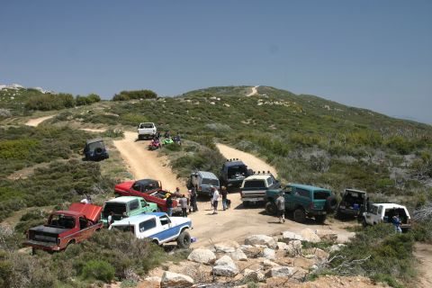 1991 Ford Bronco - Several off-road vehicles are gathered on a dusty trail in a dry, hilly terrain under a clear blue sky.
