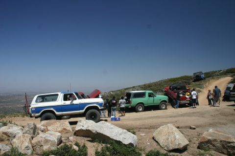 1991 Ford Bronco - A group of off-road vehicles, including a blue and white Ford Bronco, gather on a dusty trail under a clear blue sky.