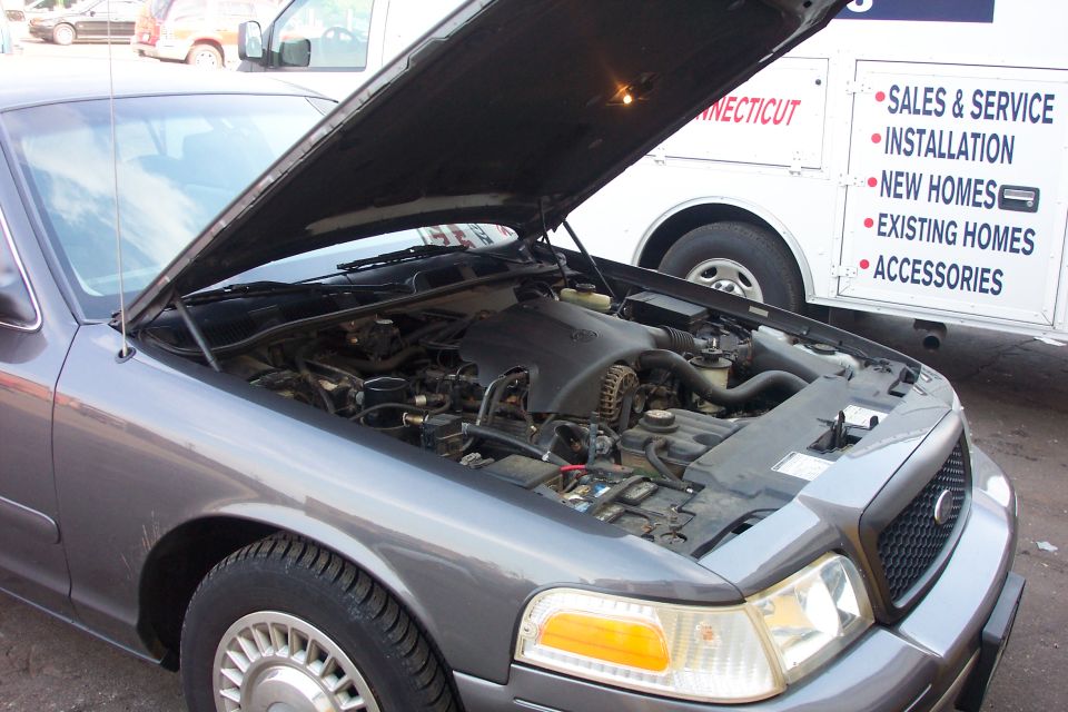 Gray 1998 Ford Crown Victoria with hood open, showing engine bay. White truck with text is behind